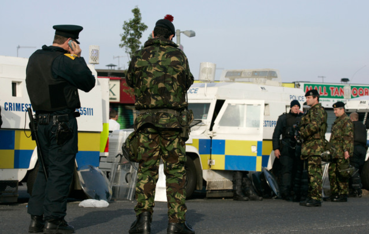 A PSNI officer on a mobile phone, and a British soldier stand in the foreground. A mixture of PSNI officers and British army soldiers stand in the background, beside a line of PSNI Landrovers.